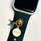 May include: A black watch band with a gold charm that says 'Mom' and has small blue and purple crystals attached to it.