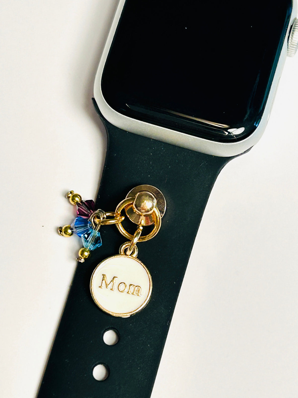May include: A black watch band with a gold charm that says 'Mom' and has small blue and purple crystals attached to it.